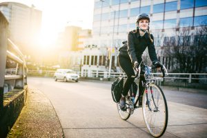 A smiling young woman commuting in an urban city environment on her street bicycle, waterproof panniers on her bike rack. She rides up a ramp from downtown Portland, Oregon, that leads up to the Burnside bridge. Colorful sky from the setting sun. Horizontal image with copy space.